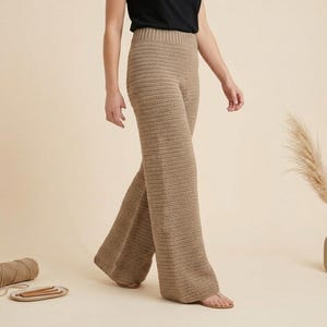 Può includere: Pantaloni all'uncinetto color beige con gamba larga. I pantaloni sono realizzati in un filato spesso e strutturato. Una matassa di filo e un piccolo vassoio con ferri da maglia sono sul pavimento. Un vaso con una pianta secca è sullo sfondo.