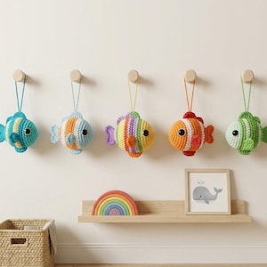 Puede incluir: Cinco adornos de peces de ganchillo de colores, colgados de clavijas de madera. Los peces presentan rayas y patrones en tonos azules, naranjas, verdes y arcoíris. Cada uno está colgado por una cuerda a juego.