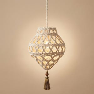 May include: A cream-colored crocheted pendant light fixture with an intricate open-weave pattern. The lamp hangs from a braided cord and features a decorative wooden bead and a matching tassel dangling from the bottom point of the structure.