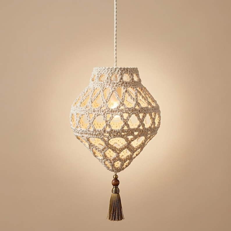 May include: A cream-colored crocheted pendant light fixture with an intricate open-weave pattern. The lamp hangs from a braided cord and features a decorative wooden bead and a matching tassel dangling from the bottom point of the structure.