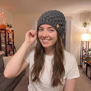 May include: A dark gray crocheted beanie with a textured bobble stitch pattern and a ribbed brim. A small, square, light brown leather-like tag is attached to the side of the beanie, featuring a subtle embossed logo design.