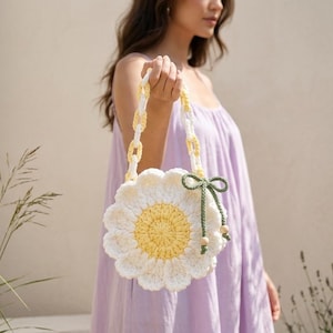 May include: A round, crocheted handbag shaped like a daisy flower with white petals and a yellow center. The bag features a handle made of translucent yellow and white plastic chain links and a dark green cord tie with small wooden beads.