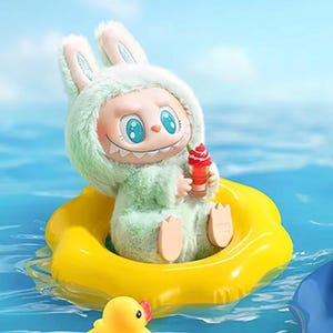 Puede incluir: Una figura caprichosa de un personaje parecido a un conejo con un atuendo verde difuso, sosteniendo un helado rojo. La figura est&aacute; sentada en un flotador inflable amarillo en el agua. Un patito de goma amarillo est&aacute; en primer plano.