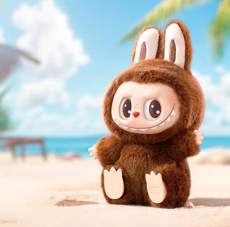 Puede incluir: Un juguete de peluche marr&oacute;n caprichoso con forma de criatura parecida a un conejo, con ojos grandes, una peque&ntilde;a nariz roja y una sonrisa. El juguete est&aacute; sobre una playa de arena con un oc&eacute;ano azul y cielo al fondo.