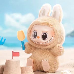Puede incluir: Un juguete de peluche con forma de conejito que lleva una capucha peluda, sosteniendo una pala amarilla. El juguete tiene una cara sonriente con ojos grandes y una hilera de dientes. Un castillo de arena con una bandera roja en primer plano.