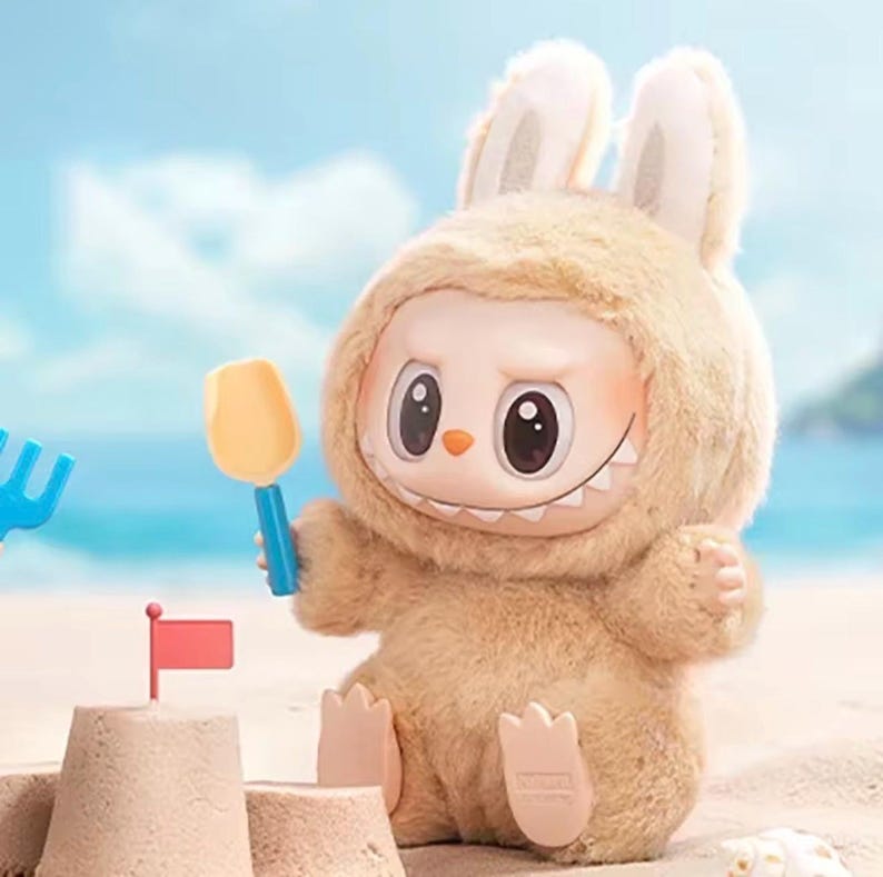 Puede incluir: Un juguete de peluche con forma de conejito que lleva una capucha peluda, sosteniendo una pala amarilla. El juguete tiene una cara sonriente con ojos grandes y una hilera de dientes. Un castillo de arena con una bandera roja en primer plano.