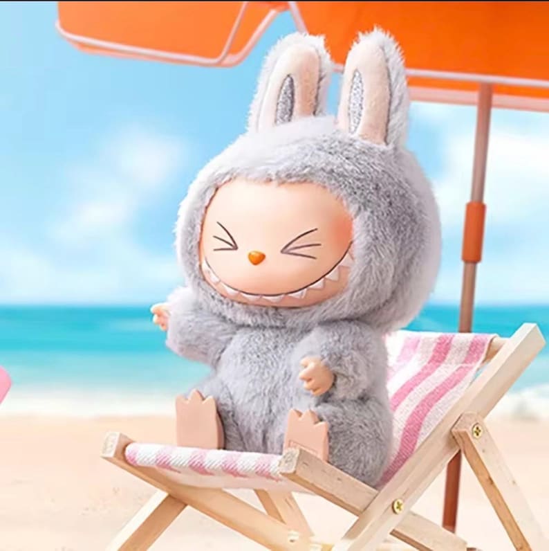 Puede incluir: Una figura caprichosa de un personaje con un disfraz de conejito gris con una cara sonriente, sentado en una silla de playa en miniatura. La escena incluye una sombrilla y una playa de arena con el oc&eacute;ano de fondo.