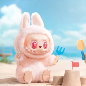 Puede incluir: Una figura de conejo rosa claro con una expresi&oacute;n traviesa, sosteniendo un rastrillo azul. La figura tiene grandes ojos rosas y una sonrisa dentuda. Est&aacute; sentada en una playa de arena con un castillo de arena y una peque&ntilde;a bandera.
