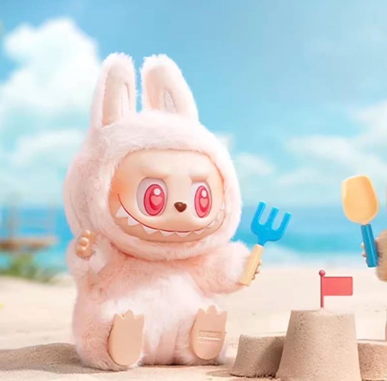 Puede incluir: Una figura de conejo rosa claro con una expresi&oacute;n traviesa, sosteniendo un rastrillo azul. La figura tiene grandes ojos rosas y una sonrisa dentuda. Est&aacute; sentada en una playa de arena con un castillo de arena y una peque&ntilde;a bandera.
