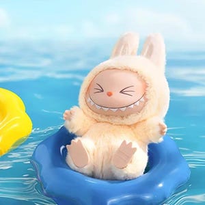 Puede incluir: Un peluche con forma de conejito que lleva un traje con capucha y una cara sonriente. El juguete es de color amarillo claro y est&aacute; en un flotador inflable azul, con un objeto inflable amarillo en el fondo. El agua es azul.