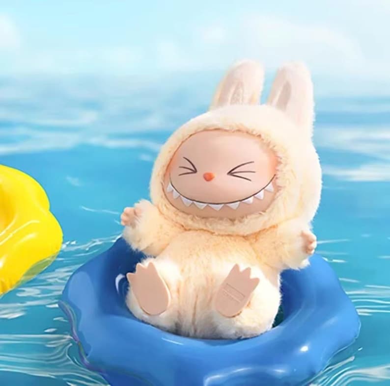 Puede incluir: Un peluche con forma de conejito que lleva un traje con capucha y una cara sonriente. El juguete es de color amarillo claro y est&aacute; en un flotador inflable azul, con un objeto inflable amarillo en el fondo. El agua es azul.