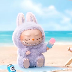 Puede incluir: Una figura caprichosa de un personaje parecido a un conejo con un traje de lavanda esponjoso, sosteniendo un helado. La figura tiene los ojos cerrados y una amplia sonrisa. Una lata de "Puck Water" est&aacute; cerca en la arena, con una playa y un oc&eacute;ano de fondo.