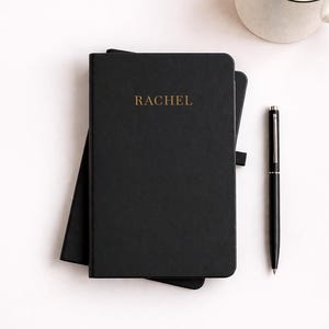 May include: Black notebook with the name "RACHEL" in gold lettering. A black pen and a mug with a yellow liquid are on the white surface. The notebook is closed and has a black ribbon bookmark.