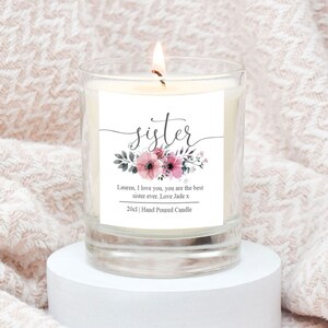May include: A clear glass candle jar with a white label that says "sister" in a cursive font. The label also has a floral design with pink and grey flowers. The text on the label reads "Lauren, I love you, you are the best sister ever. Love Jade x 200ml Hand Poured Candle".
