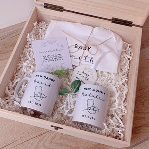 May include: A wooden gift box filled with baby shower gifts. The box contains two white mugs with the text "New Daddy" and "New Mummy" with names and the year 2024. There is also a white onesie with the text "Baby Smith" and a small round ornament with the text "Baby Smith May 2024".