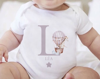 May include: A white baby onesie with a purple letter L and a hot air balloon graphic. The onesie has the name "LÉA" printed below the balloon.