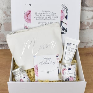 May include: A white gift box filled with a variety of items for Mother's Day. The box includes a white canvas bag with the word "Mum" written on it, a small jar of bath salts, a candle with a floral design, a small bottle of aloe vera lotion, and a silver necklace with a heart pendant. The box is decorated with a floral pattern and the words "Happy Mother's Day".