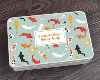 May include: A silver metal tin with a blue background and a pattern of orange, red, and black fish. The text "Grandad George's Fishing Things" is printed on the tin.