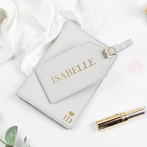 May include: A gray leather passport holder with a matching luggage tag. The luggage tag has the name "Isabelle" printed in gold lettering. The passport holder has a heart with the letters "ID" printed in gold.