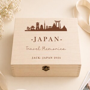 May include: A light-colored wooden box with a hinged lid. The lid features a brown silhouette of Japanese landmarks, the word "-JAPAN-", and the text "Travel Memories". The box is secured with a metal clasp and includes the text "JACK - JAPAN 2024".