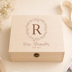 May include: A light-coloured wooden box with a hinged lid. The lid features a brown floral wreath design with the letter "R" in the centre. Below the wreath is the name "Ross Alexander" and the date "28.07.2021". A metal clasp secures the box.