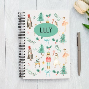 Pode incluir: Um caderno branco de espiral com um design colorido que apresenta um quebra-nozes, uma bailarina e uma árvore de Natal. O caderno tem um oval verde com o nome "LILLY" escrito em branco.
