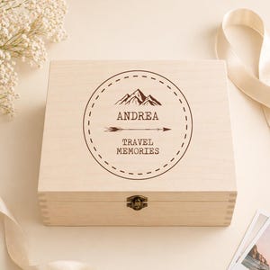 May include: A light-coloured wooden box with a hinged lid and a metal clasp. The box is decorated with a circular design featuring a mountain range, an arrow, and the text "ANDREA TRAVEL MEMORIES". Decorative elements surround the box.