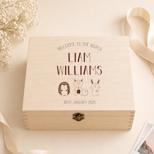 May include: A light-colored wooden keepsake box with the text "WELCOME TO THE WORLD" and the name "LIAM WILLIAMS" in brown lettering. The box features illustrations of a superhero, a fox, and a rabbit. The date "30TH JANUARY 2021" is also printed on the box.