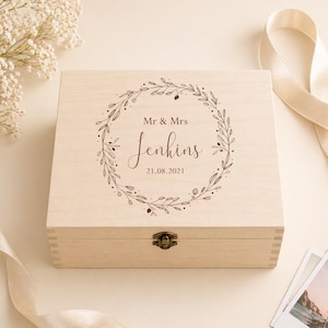 May include: A light-colored wooden box with a hinged lid and a metal clasp. The box is engraved with the words "Mr & Mrs Jenkins" and the date "21.08.2021" inside a decorative wreath. The box is surrounded by flowers and a ribbon.