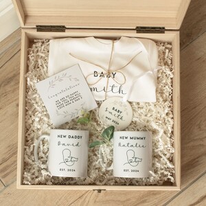 May include: A wooden box filled with baby shower gifts, including a white onesie with the text "Baby Smith", a white mug with the text "New Daddy David Est. 2024", a white mug with the text "New Mummy Natalie Est. 2024", a white ornament with the text "Baby Smith May 2024", and a card with the text "Congratulations We are so happy for you. You will make fab parents. Love Mum & Dad x".
