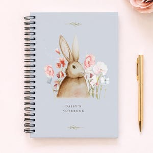 May include: A spiral-bound notebook with a light blue cover featuring a watercolor illustration of a brown rabbit surrounded by pink and white flowers. The text "DAISY'S NOTEBOOK" is printed in black below the rabbit. A gold pen lies to the right.