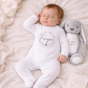 Puede incluir: Un beb&eacute; durmiendo con un body blanco con el nombre "Freya" en un dise&ntilde;o de corona floral. Un conejito de peluche gris con "Freya" en la oreja descansa junto al beb&eacute;. Tambi&eacute;n hay una tarjeta "Hello Baby".
