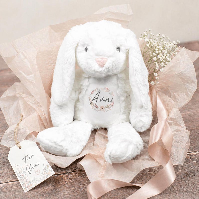 Puede incluir: Un conejo de peluche blanco con orejas largas, una nariz rosa y el nombre "Ava" en una corona floral. El conejo est&aacute; envuelto en papel de seda rosa con una etiqueta de regalo que dice "For You". Una ramita de flores blancas y una cinta rosa completan la presentaci&oacute;n.