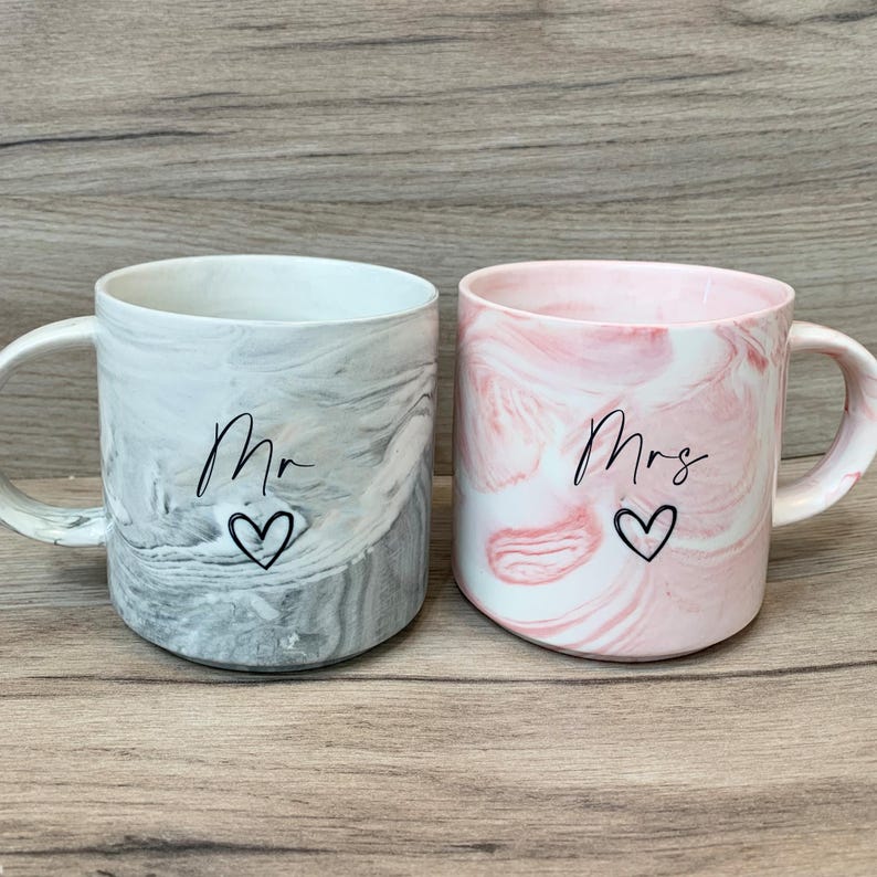 Puede incluir: Dos tazas de cer&aacute;mica con dise&ntilde;o de m&aacute;rmol. Una taza es gris con la palabra "Mr." y un coraz&oacute;n. La otra taza es rosa con la palabra "Mrs." y un coraz&oacute;n.