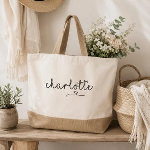 May include: A cream-colored canvas tote bag with a tan burlap base. The front of the bag features the name Charlotte printed in black script lettering, accompanied by a small heart icon centered below the name.