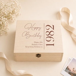May include: A wooden birthday box with a hinged lid. The lid features the words "Happy Birthday" in a script font, the year "1982", and a personalized message. A metal clasp secures the box. Decorative elements include flowers and a ribbon.