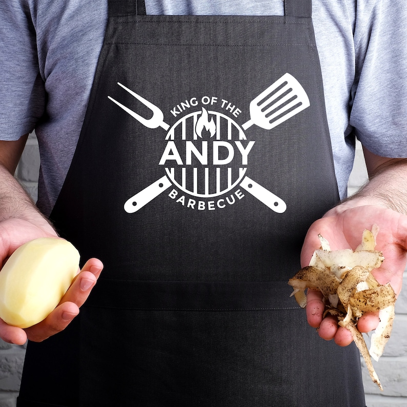 May include: A person wearing a black apron with a white logo that says "King of the Andy Barbecue". The person is holding a peeled potato in one hand and potato peelings in the other.