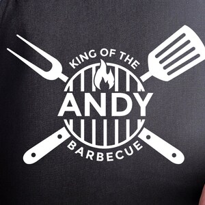 May include: White text on a black background reads "King of the BBQ" with a grill graphic and flames. The text "ANDY" is in the centre of the grill graphic. A spatula and a fork are crossed behind the grill graphic.