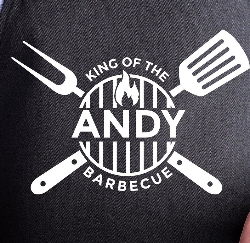May include: White text on a black background reads "King of the BBQ" with a grill graphic and flames. The text "ANDY" is in the centre of the grill graphic. A spatula and a fork are crossed behind the grill graphic.