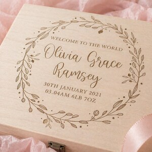 May include: A light-colored wooden box with engraved text and a floral wreath design. The text reads "WELCOME TO THE WORLD" above the name "Olivia Grace Ramsey", birth date, time, and weight: 6lb 7oz.