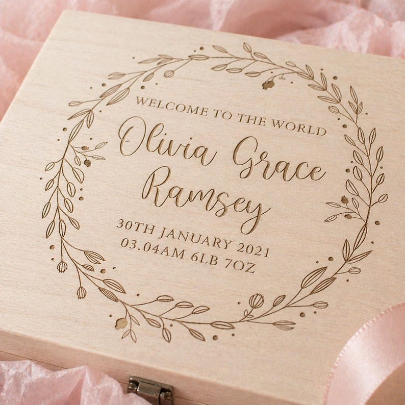 May include: A light-colored wooden box with engraved text and a floral wreath design. The text reads "WELCOME TO THE WORLD" above the name "Olivia Grace Ramsey", birth date, time, and weight: 6lb 7oz.