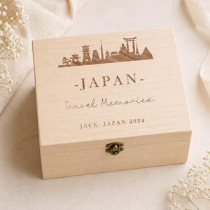 May include: A light-colored wooden box with a decorative lid featuring a silhouette of Japanese landmarks. The lid is engraved with the word "JAPAN", "Travel Memories", and "JACK- JAPAN 2024". The box has a metal clasp.