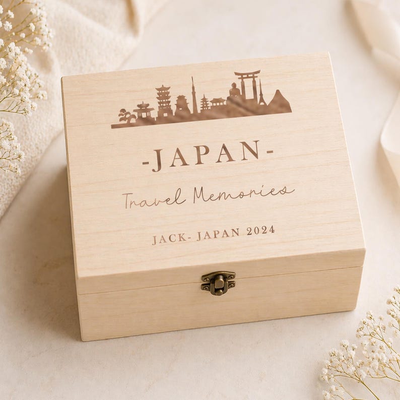 May include: A light-colored wooden box with a decorative lid featuring a silhouette of Japanese landmarks. The lid is engraved with the word "JAPAN", "Travel Memories", and "JACK- JAPAN 2024". The box has a metal clasp.
