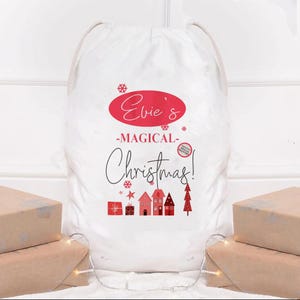 May include: A white Christmas gift sack with a drawstring closure. The sack features the text "Evie's -MAGICAL- Christmas!" in red and black, along with red illustrations of gifts, houses, and a Christmas tree. Presents wrapped in brown paper are in the foreground.