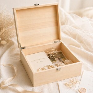 May include: An open, light-colored wooden box with a hinged lid. Inside, a wedding photo, a booklet that says "Our Wedding", and a strand of pearls are visible. The box is on a white surface.