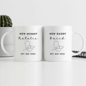 May include: Two white ceramic mugs with black text and a simple line drawing of a baby in the womb. The mug on the left says "New Mummy Natalie Est. May 2024". The mug on the right says "New Daddy David Est. May 2024".