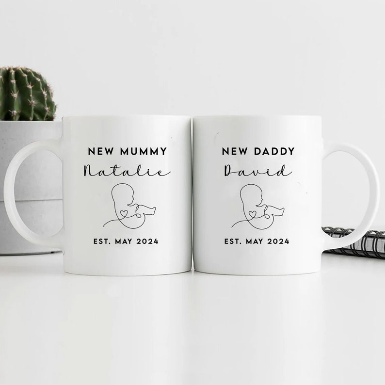 May include: Two white ceramic mugs with black text and a simple line drawing of a baby in the womb. The mug on the left says "New Mummy Natalie Est. May 2024". The mug on the right says "New Daddy David Est. May 2024".