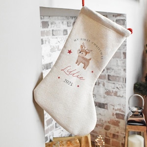 May include: A cream Christmas stocking with a red hanging loop. It features a cartoon reindeer illustration, the text "MY FIRST CHRISTMAS", the name "Lillie", and the year "2025" in red.