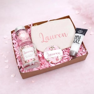 May include: A gift box filled with pink paper shreds containing a beige canvas pouch with the name Lauren in pink, a glass candle jar labeled Lauren Will You Be My Bridesmaid, a round floral mirror with the name Lauren, and a tube of Natural lotion.