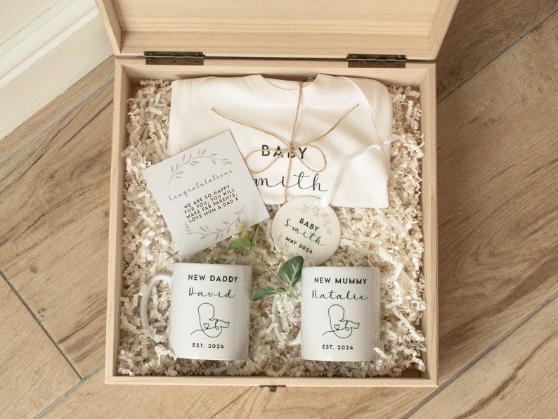 May include: A wooden box filled with baby shower gifts, including a white onesie with the text "Baby Smith", a white mug with the text "New Daddy David Est. 2024", a white mug with the text "New Mummy Natalie Est. 2024", a white ornament with the text "Baby Smith May 2024", and a card with the text "Congratulations We are so happy for you. You will make fab parents. Love Mum & Dad x".